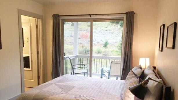 A bedroom with a bed, a nightstand, a lamp, and a view of a balcony with chairs. - Big Sky Haven, Quiet and Centrally Located for Skiing, Fishing, Golfing 
 (Big Sky)