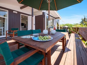 Een houten terras met een eettafel, groene bekleden stoelen en een grote parasol.