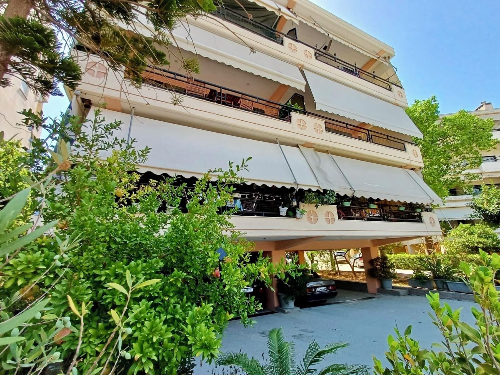 Un edificio de varios pisos con balcones, plantas en macetas y un coche estacionado en la entrada.