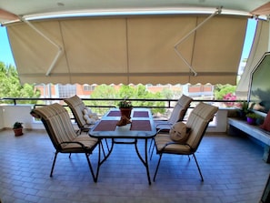 A covered patio with a table and four chairs, potted plants, and a view of buildings and trees.