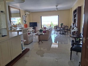 A spacious living area with a sofa, coffee table, and dining table. There is a television and a large window providing natural light.