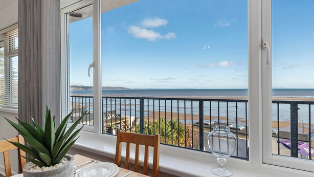 A balcony with a view of the beach, a dining table with plates and a glass, and a potted plant.