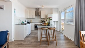 A modern kitchen with white cabinets, a black countertop, wooden stools, and a large window with blinds.