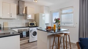 A compact kitchen with a microwave, oven, and washing machine, a small island with stools, and a window with blinds.