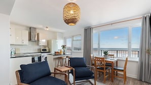 A modern kitchen with white cabinets, a stainless steel range hood, and a dining area with wooden furniture and a view of the sea.