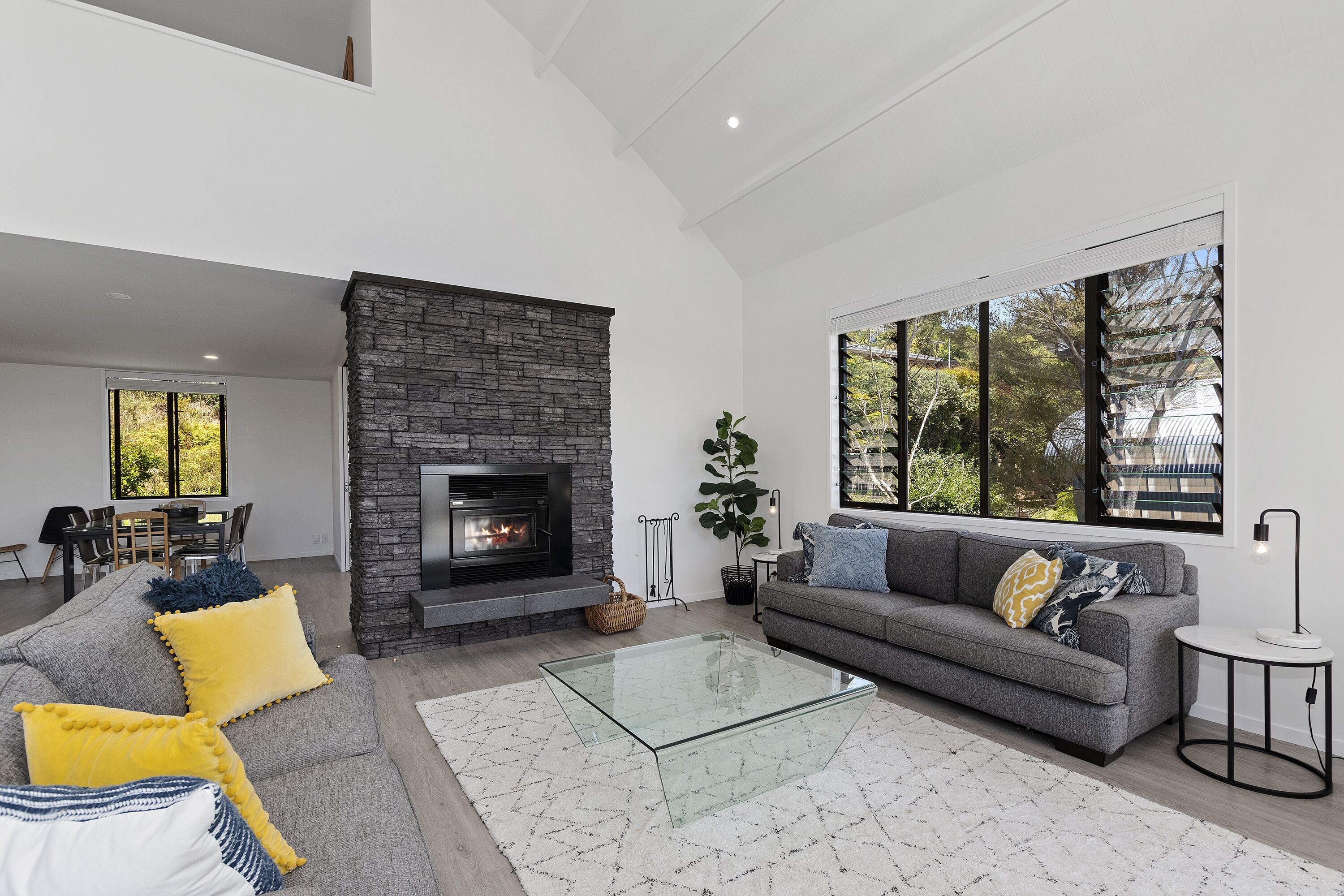 A modern living room with a fireplace, a grey sofa, yellow and white cushions, a glass coffee table, and a large window with a view of trees.