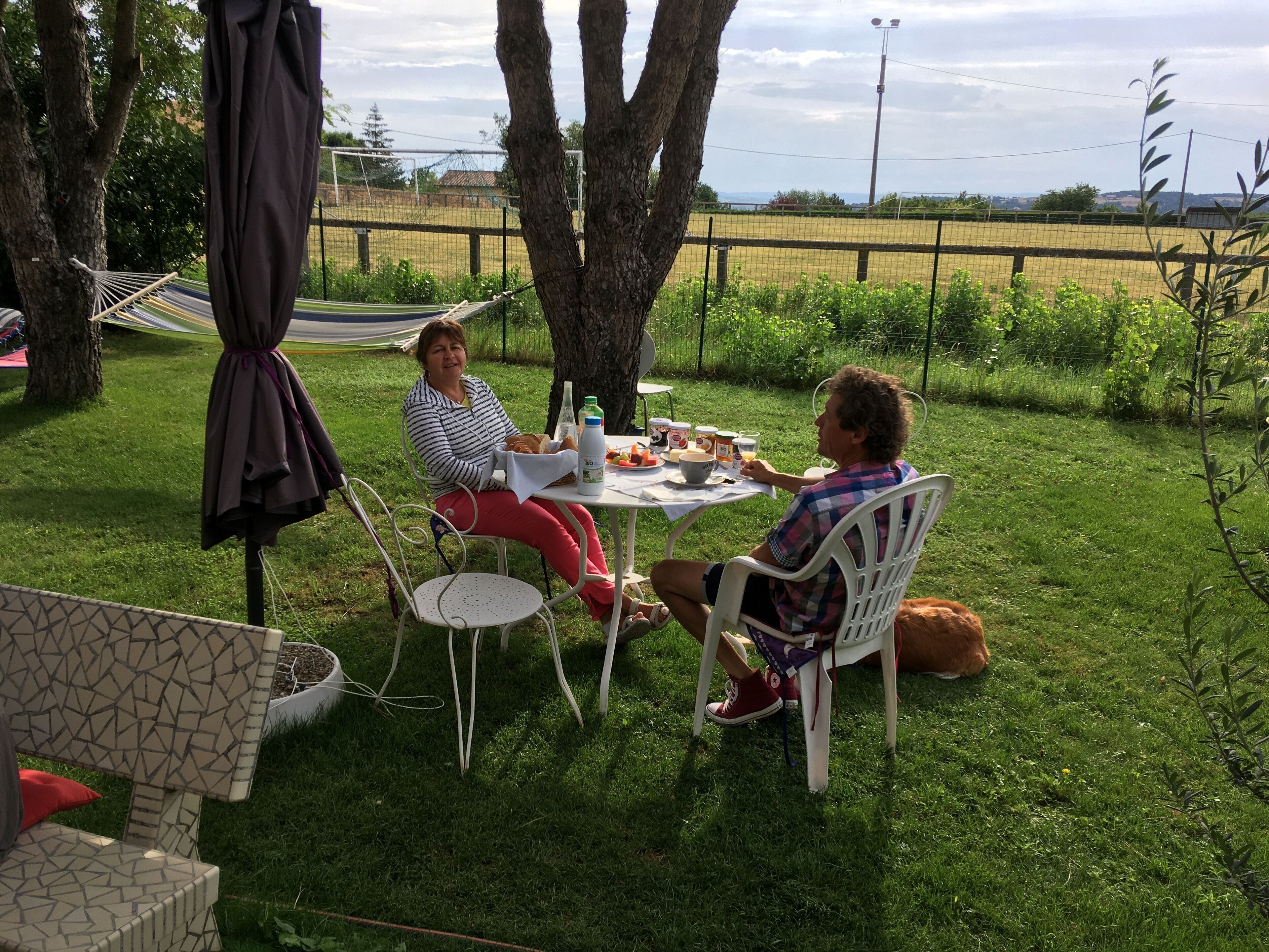Two people seated at an outdoor table with a picnic setup, surrounded by greenery and trees.
