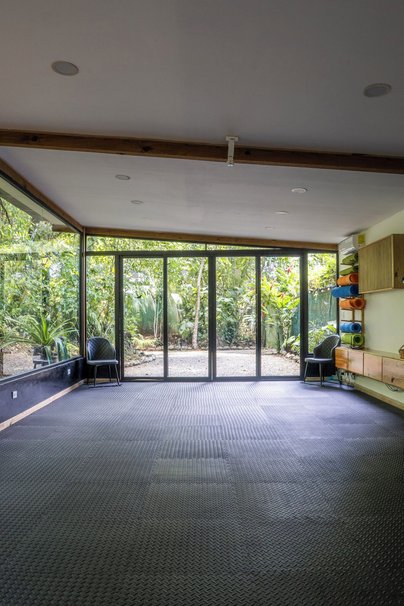 A spacious room with large sliding glass doors, a black cushioned seating area, and a wall-mounted shelf with rolled-up mats.