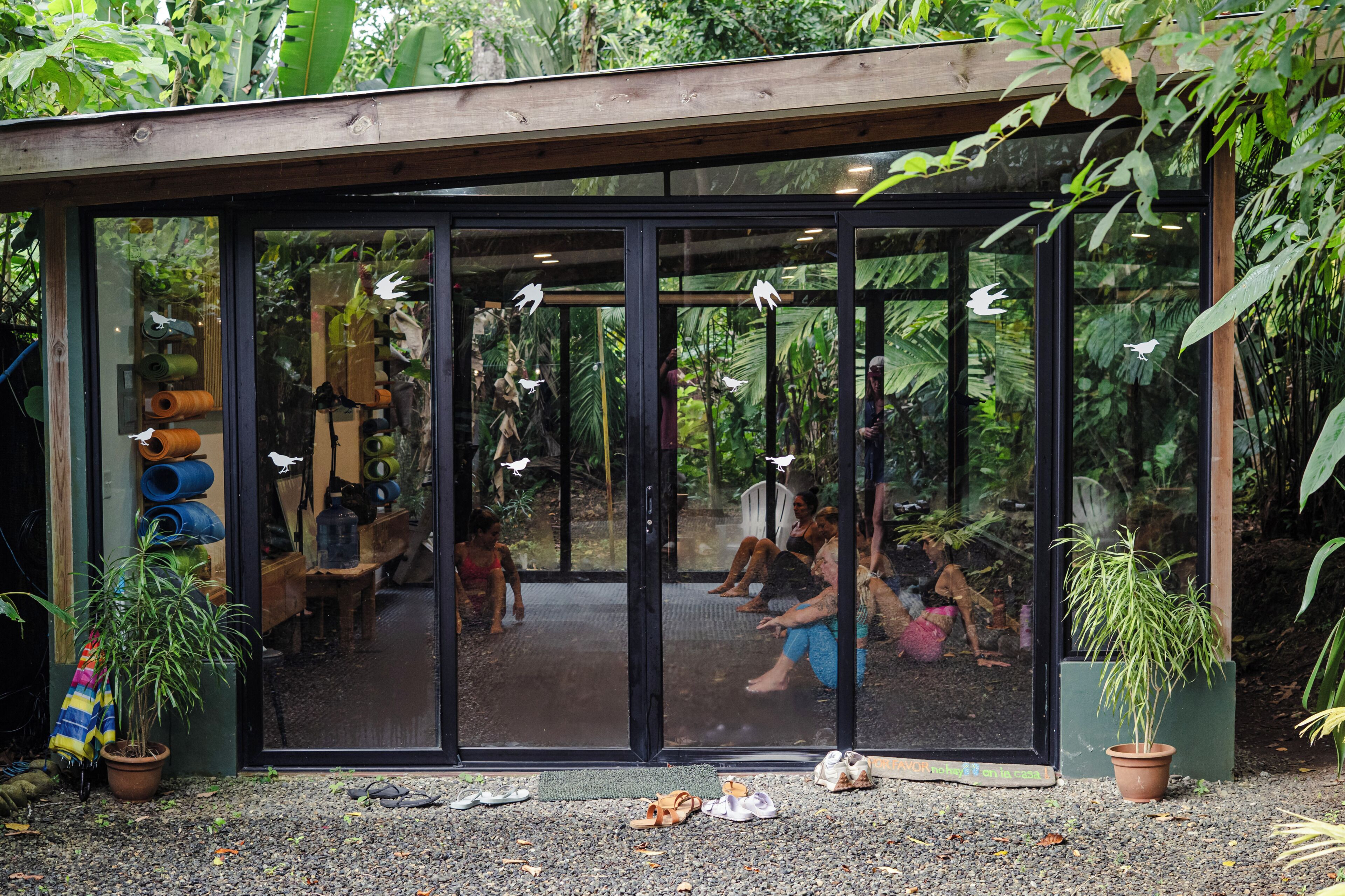 A glass-enclosed structure with a wooden roof, surrounded by greenery. Inside, there are people seated on the floor, and various items are visible through the glass.