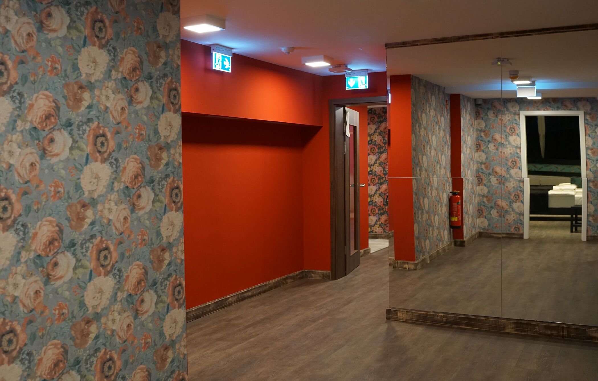 Ein Hotelgang mit blumengemusterten Tapeten, roten Wänden und einer Spiegelwand, die den Raum reflektiert.