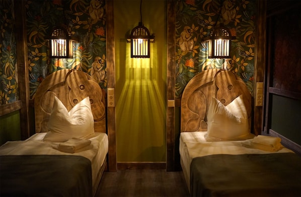 Una habitación con dos camas, cada una con cabecero de madera y almohada blanca. Hay una cortina verde en el centro, y una lámpara de pared sobre la cortina.