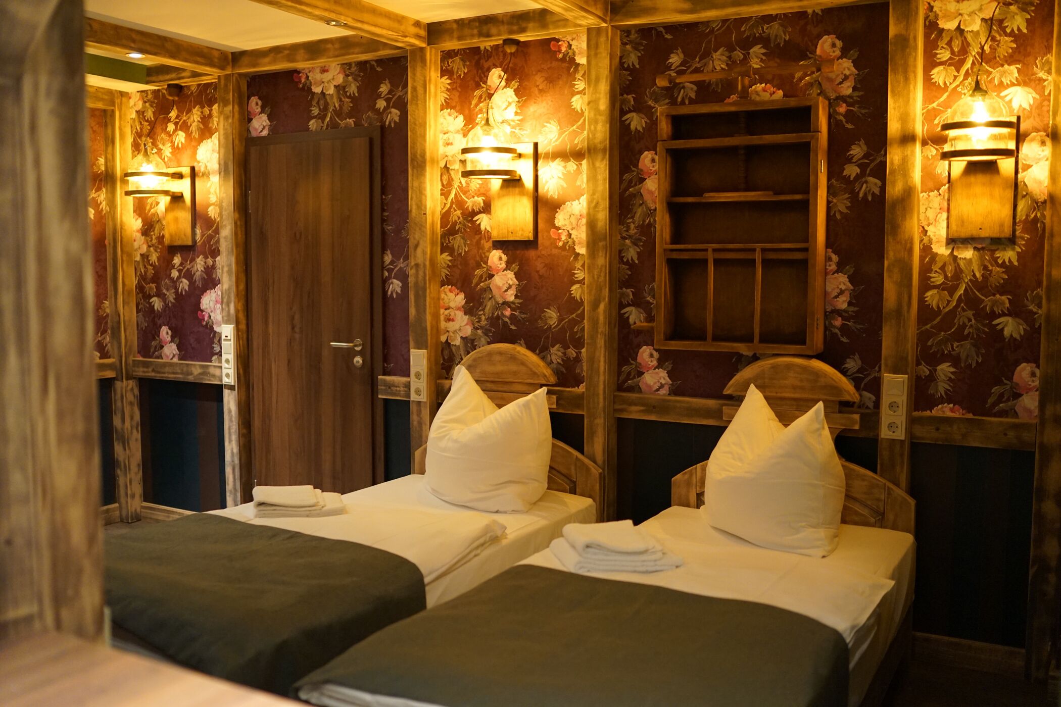 A room with two beds, each with a pillow and a folded towel. There are wall-mounted light fixtures and a wooden door.
