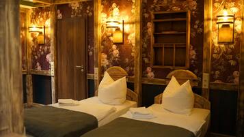 A room with two beds, each with a pillow and a folded towel. There are wall-mounted light fixtures and a wooden door.