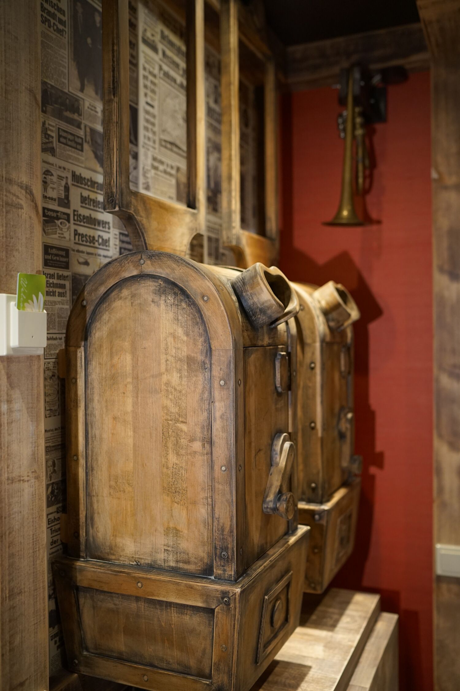 A wooden cabinet with a brass bell, set against a red wall with newspaper clippings.