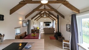A spacious living area with exposed wooden beams, a dining table with a red tablecloth, a kitchen area with a stove, and a large window with curtains. - The Dairy - Luxury Cottage with Hot Tub and Views (Ludchurch)