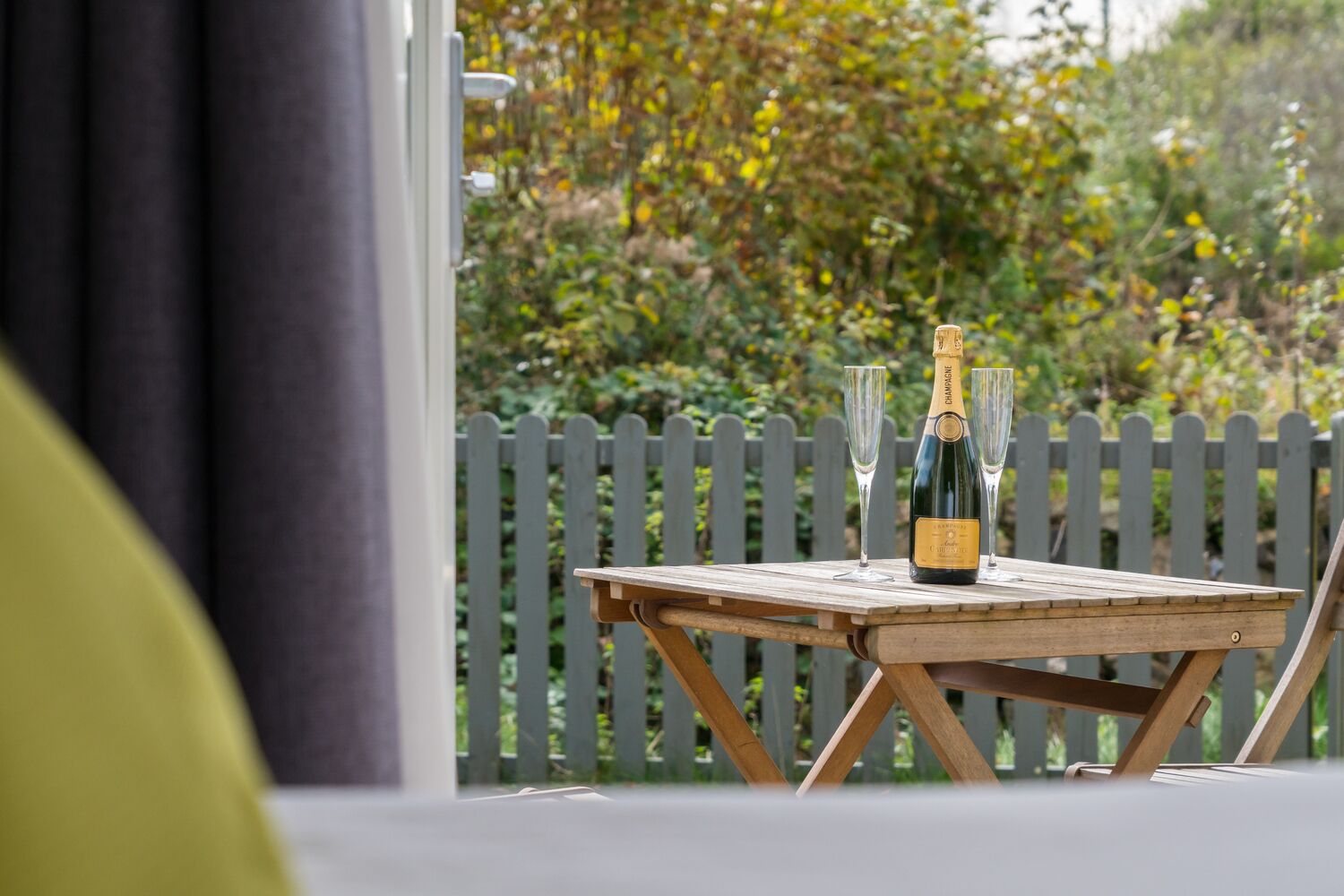 Un tavolo di legno con una bottiglia di champagne e due bicchieri, con sullo sfondo una recinzione in legno e vegetazione.