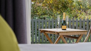 A wooden table with a bottle of champagne and two glasses on it, set against a backdrop of a wooden fence and greenery. - The Dairy - Luxury Cottage with Hot Tub and Views (Ludchurch)