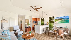 A spacious living area with a sofa, coffee table, and dining set. A kitchen with wooden cabinets and a bar area is visible. - KIAHUNA BEACH HOUSE #50 NEW REMODEL GROUND FLOOR NEW AC TWIN BRIDGES GARDEN VIEW (Koloa)