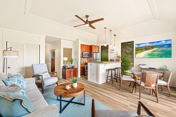 A spacious living area with a sofa, coffee table, and dining set. A kitchen with wooden cabinets and a bar area is visible.
