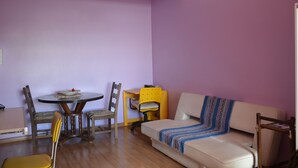 Une pièce avec une table de salle à manger ronde, deux chaises, un bureau jaune, un canapé beige et un mur violet.
