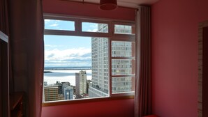 Une chambre avec une grande fenêtre, un lit et une vue sur le paysage urbain et l’étendue d’eau.
