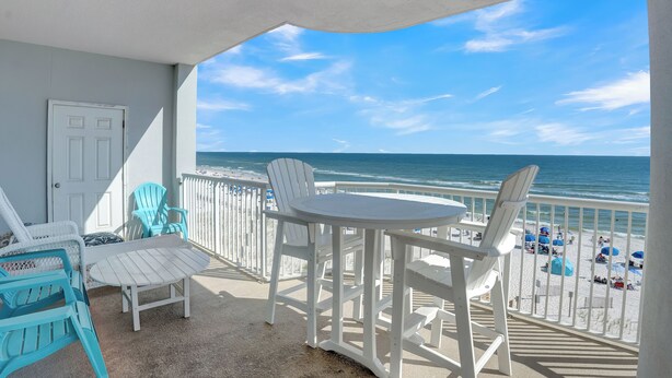 Un balcon avec des chaises blanches et une table, donnant sur une plage parsemée de parasols et fréquentée par des gens.