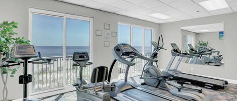 A gym with treadmills, elliptical machines, and a stationary bike, large windows with a view of the ocean, and a ceiling with recessed lighting.