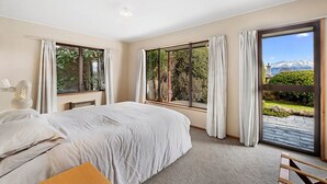 Une chambre avec un grand lit, deux fenêtres avec des rideaux, une vue sur la verdure et les montagnes, et une petite table avec une lampe.