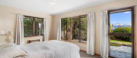 A bedroom with a large bed, two windows with curtains, a view of greenery and mountains, and a small table with a lamp.