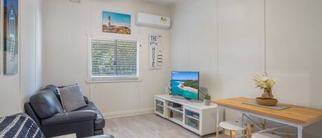 Un salon avec un canapé en cuir, un téléviseur posé sur un meuble blanc, une table en bois avec deux tabourets et une fenêtre avec des stores.