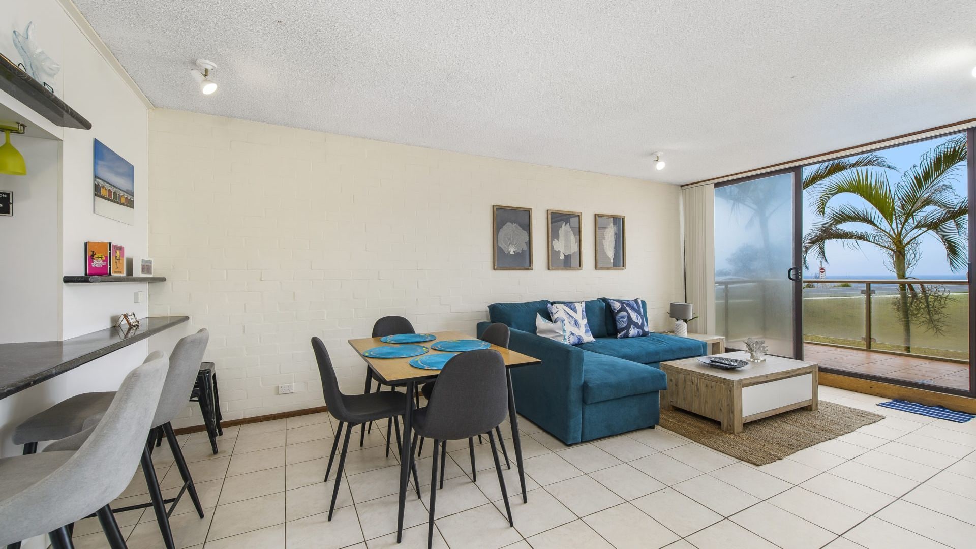 A modern living room with a blue sofa, dining table, and chairs. There are framed pictures on the wall and a view of the sea through a large window.