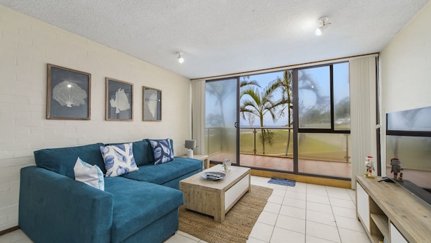 Een moderne woonkamer met een blauwgroene bank, een salontafel en een grote schuifdeur die leidt naar een balkon met uitzicht op palmbomen.