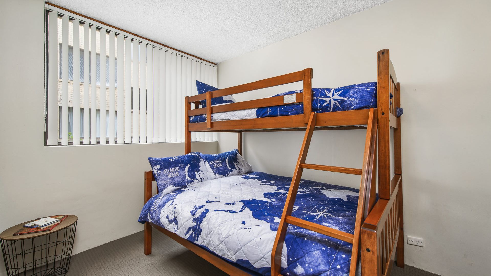 A bunk bed with a star-patterned bedspread, a window with blinds, and a small round table with books.