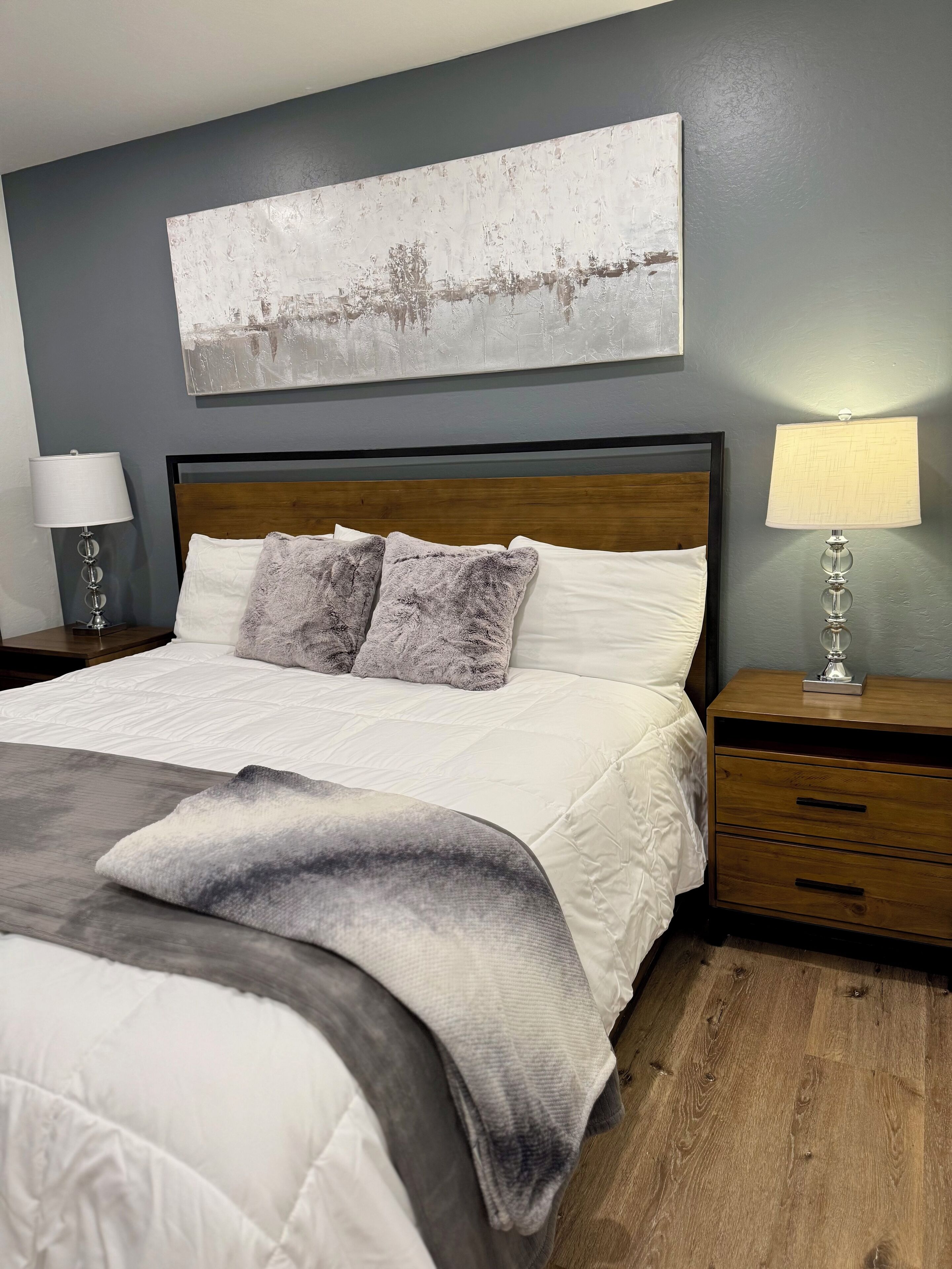 A bedroom with a large bed, two bedside tables, a nightstand, a wall-mounted lamp, and a painting above the bed.
