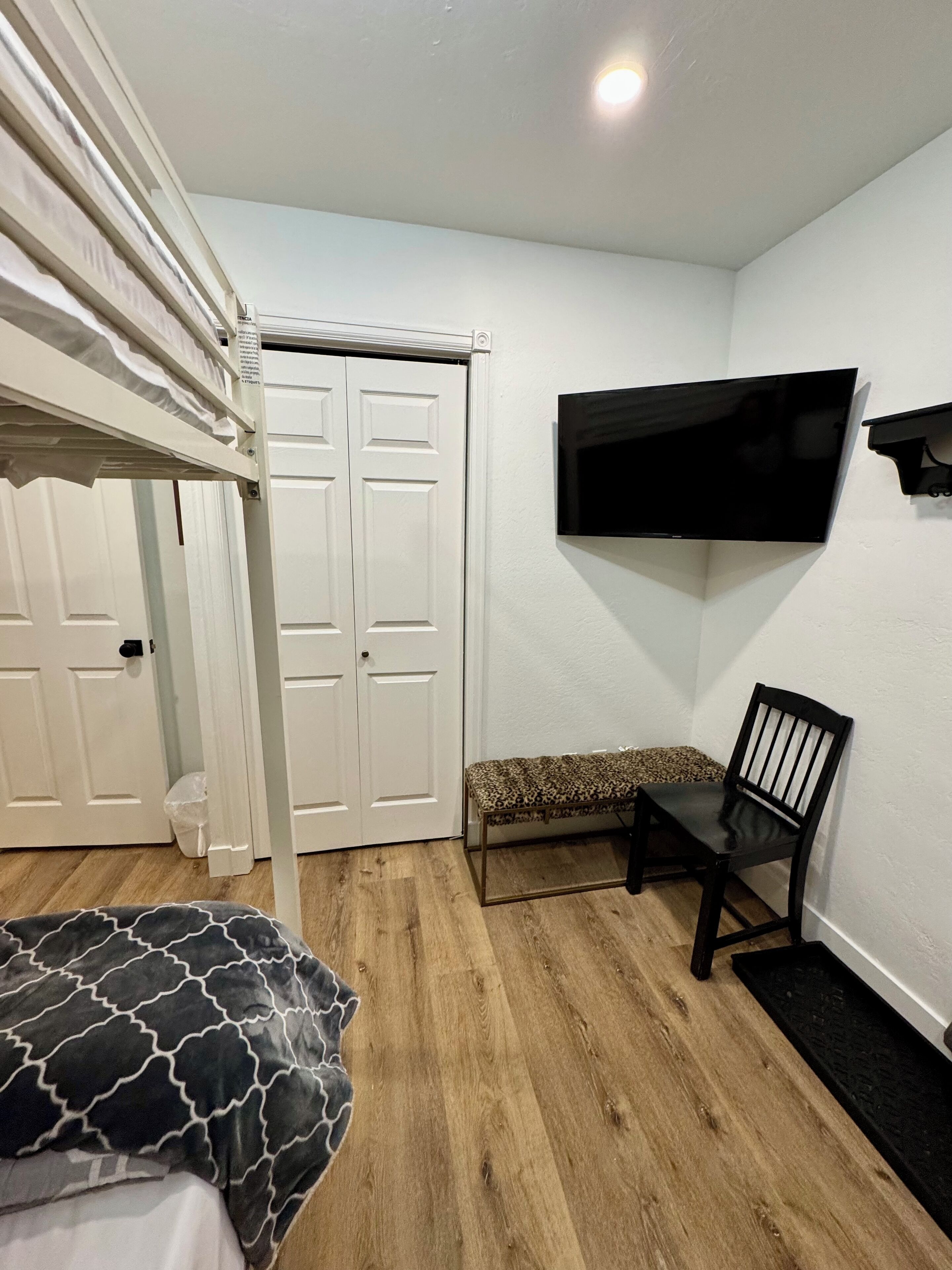A bedroom with a bunk bed, a bench, a chair, a TV mounted on the wall, and a door with a trash bin beside it.