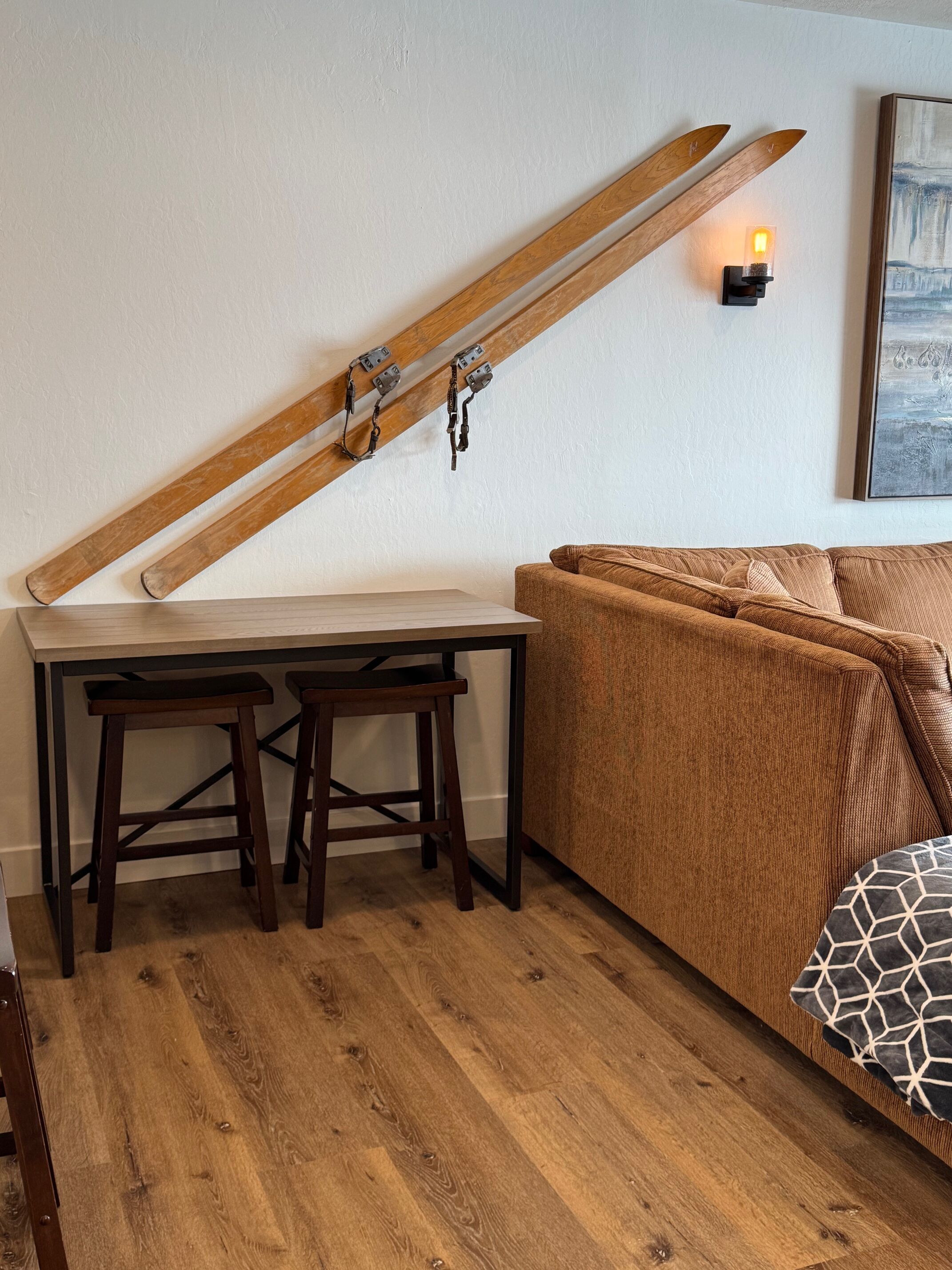A room with a wooden slat ceiling, a small table with two stools, a brown sofa, and a wall-mounted light fixture.