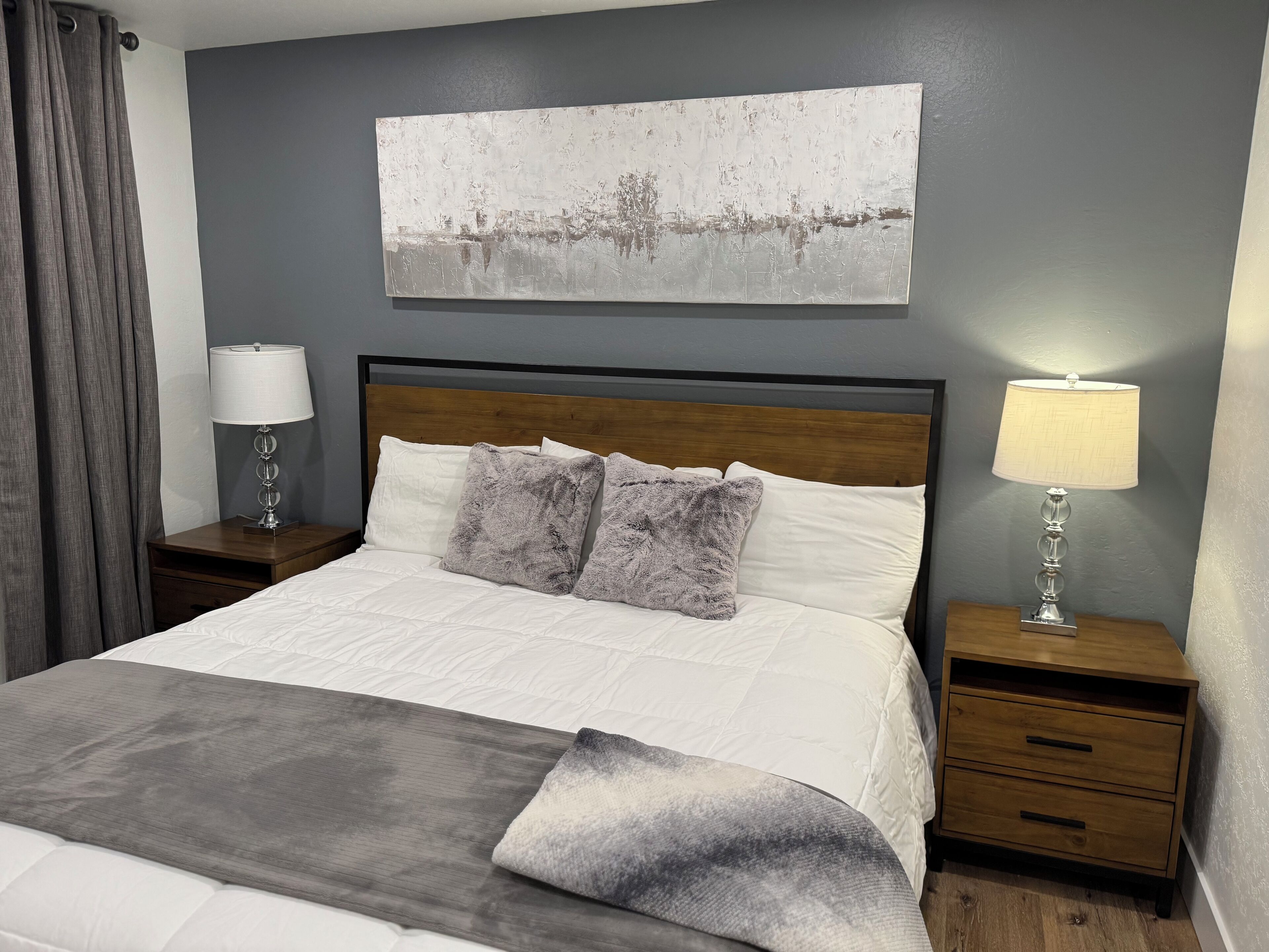 A neatly made bed with white and gray bedding, two bedside tables with lamps, a gray wall, and a painting above the bed.