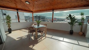 A covered outdoor area with a table and chairs, potted plants, and a view of the cityscape. - City Life (Iskenderun)