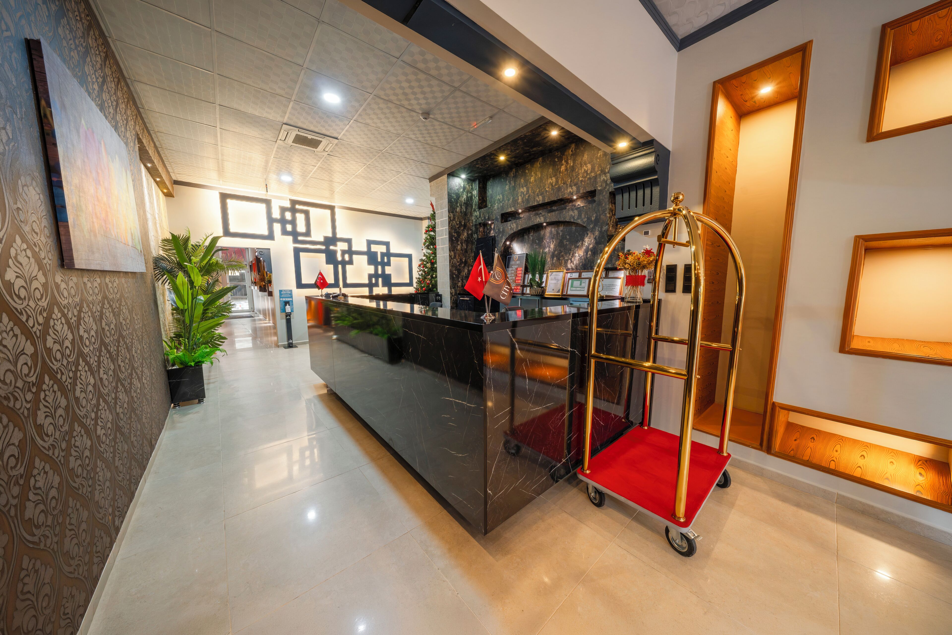 A hotel reception area with a modern design, featuring a reception desk, a red and gold cart, and a large potted plant.
