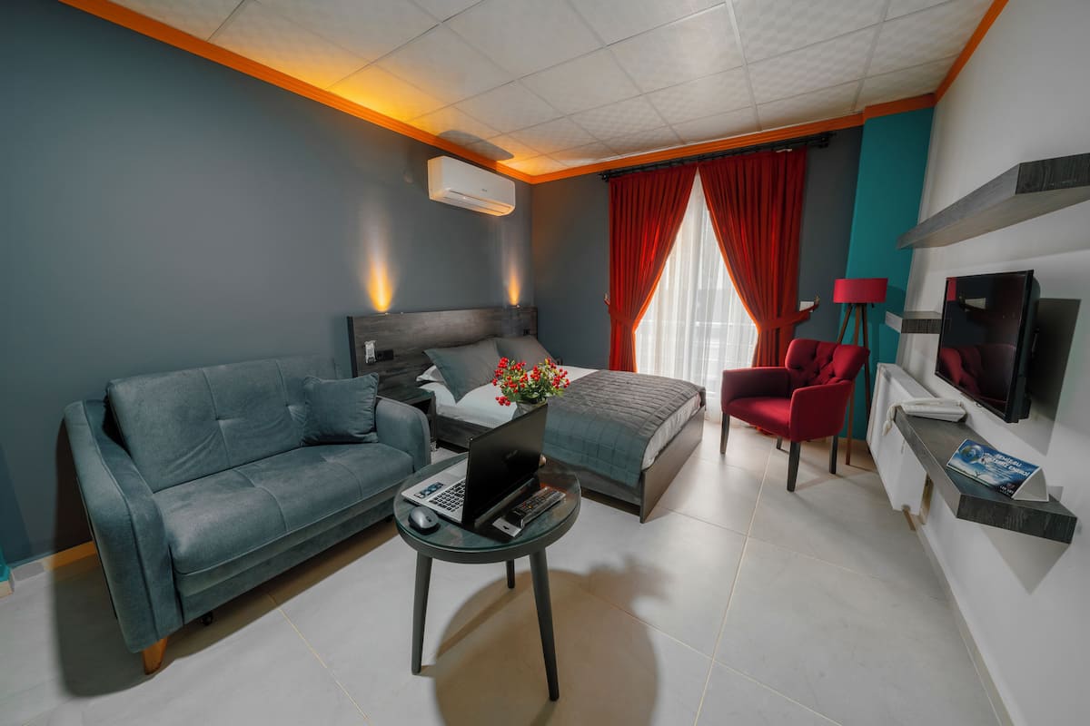A modern hotel room with a sofa, a bed, a desk with a laptop, a TV, and a red chair.