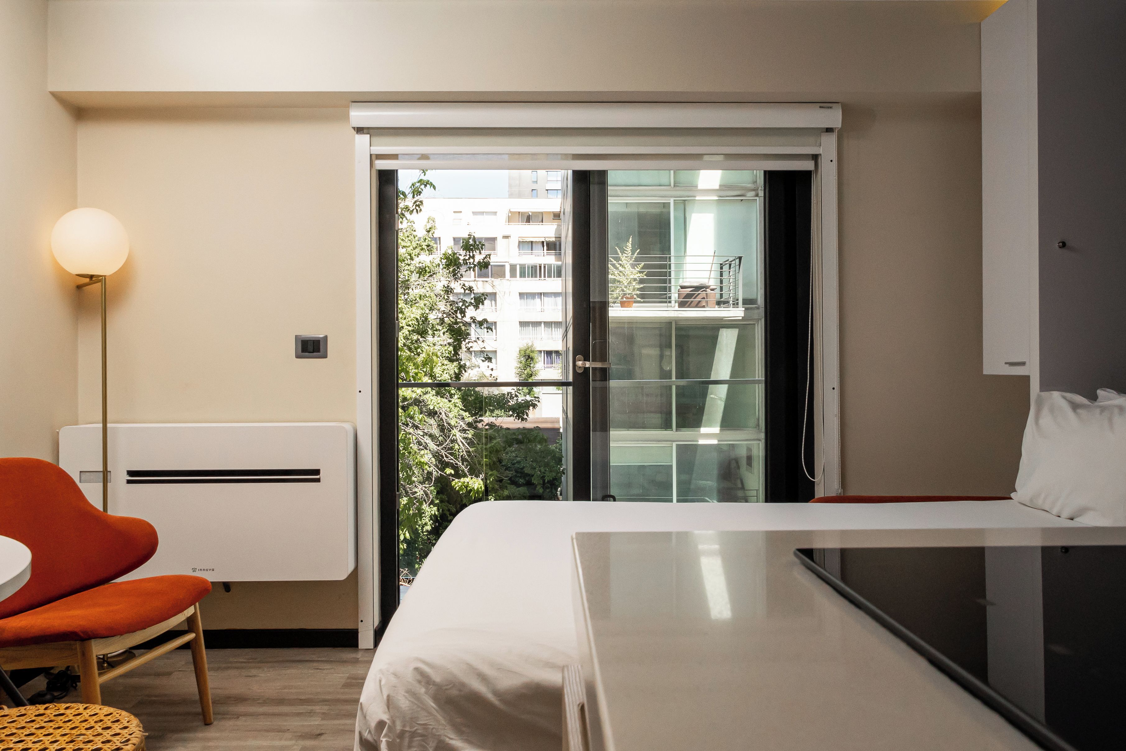 A hotel room with a bed, a chair, a table, a lamp, and a view of a building through a glass door.