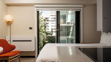 A hotel room with a bed, a chair, a table, a lamp, and a view of a building through a glass door.