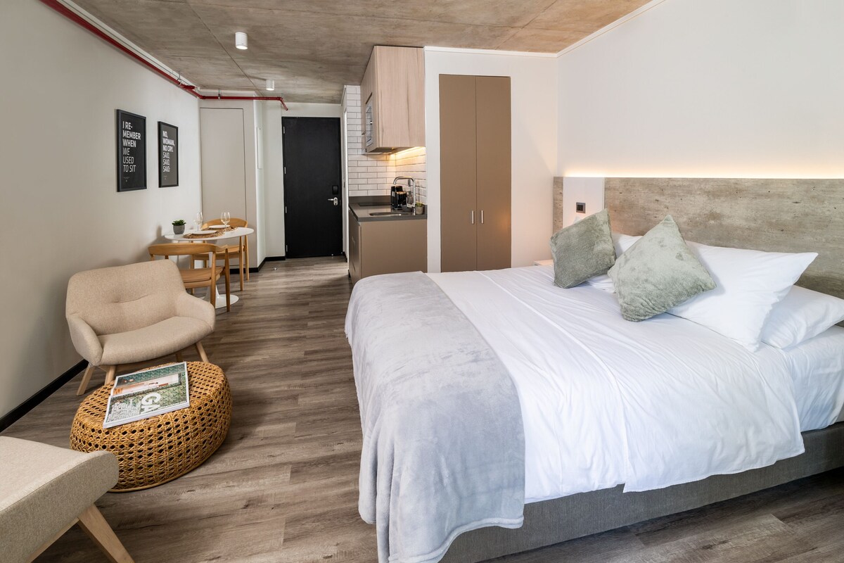 A modern hotel room with a large bed, a small kitchenette, a dining area, and a separate bathroom.
