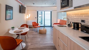 Cocina moderna con microondas, tostadora y lavabo. Hay una zona de comedor con sillas naranja y una mesa redonda. Una cita enmarcada está en la pared.
