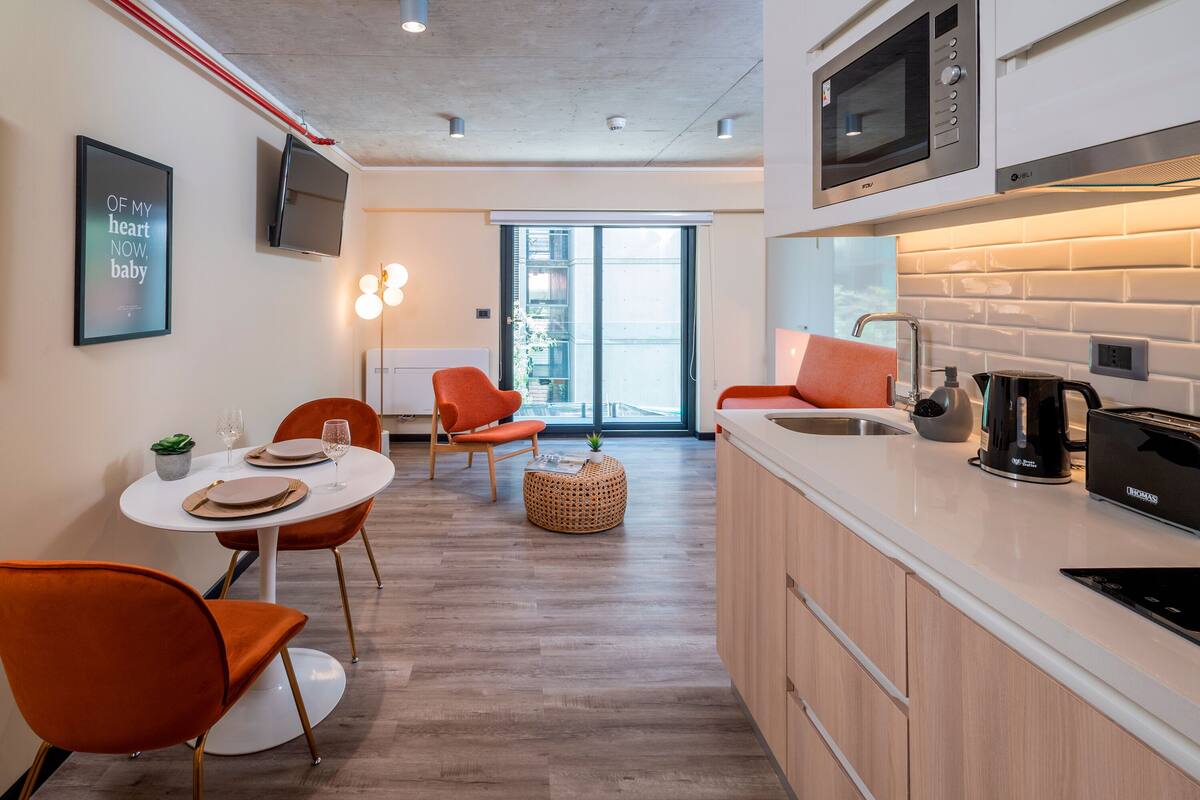 Cocina moderna con microondas, tostadora y lavabo. Hay una zona de comedor con sillas naranja y una mesa redonda. Una cita enmarcada está en la pared.