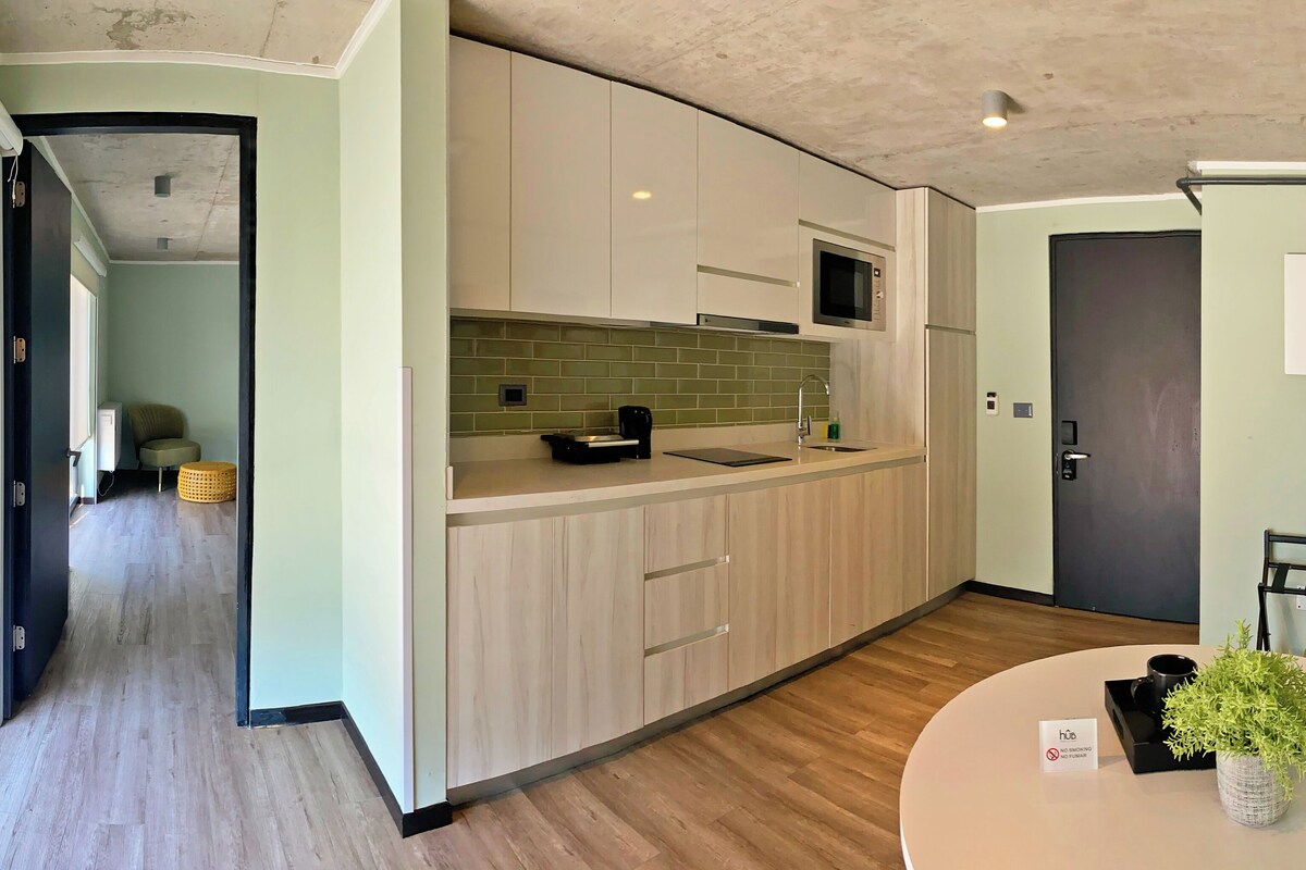 A modern kitchen with light wood cabinets, a microwave, and a sink. There is a dining area with a round table and chairs, and a small plant on the table.