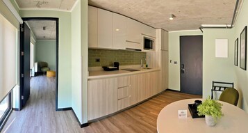 Una cocina moderna con armarios de madera clara, microondas y un lavabo. Hay una zona de comedor con una mesa redonda y sillas, y una planta pequeña sobre la mesa.