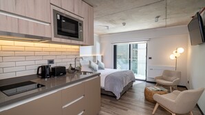 Una cocina moderna con microondas, cafetera y tostadora. Hay una cama con manta y almohadas grises, una silla y una lámpara de pie.