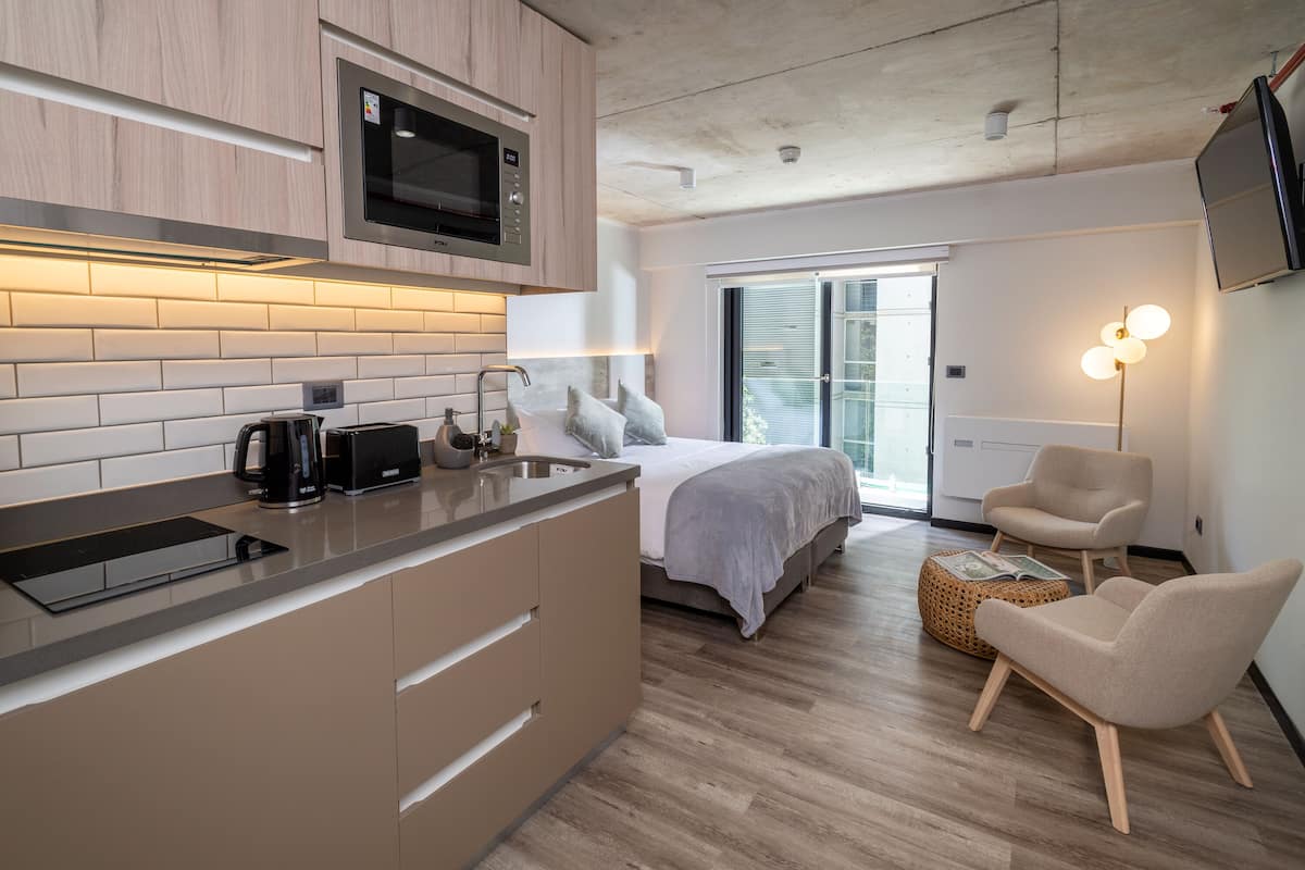 Una cocina moderna con microondas, cafetera y tostadora. Hay una cama con manta y almohadas grises, una silla y una lámpara de pie.