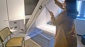 A person installing a white, wall-mounted, foldable bed in a room with a wooden floor, a chair, and a window.